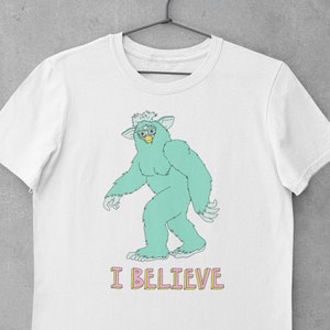 May include: White t-shirt with a cartoon green monster wearing glasses and the text "I BELIEVE" in pink and yellow letters.