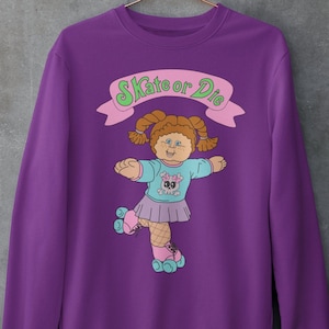 May include: Purple sweatshirt with a graphic of a Cabbage Patch Kid doll on roller skates. The doll is wearing a pink skirt, blue shirt, and fishnet stockings. The text "Skate or Die" is above the doll in a green and pink banner.
