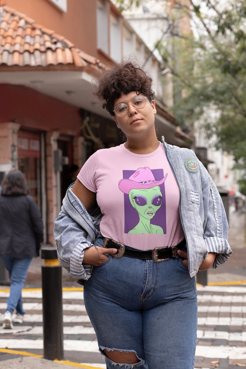 May include: A pink t-shirt with a graphic of an alien wearing a pink cowboy hat. The alien has green skin and large eyes with glitter. The shirt is being worn by a person with curly brown hair and glasses.