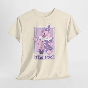 May include: A cream-colored short-sleeve t-shirt featuring a graphic of a teddy bear dressed as a clown in a purple and pink outfit with a matching hat. The bear is inside a rectangular frame with the text The Fool printed below it.