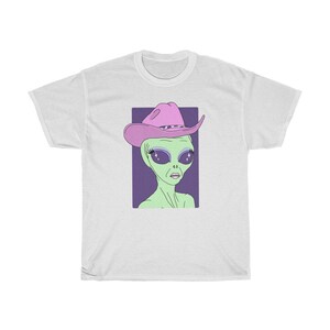 May include: A white t-shirt with a green alien wearing a pink cowboy hat. The alien has large, sparkling eyes and a purple background.