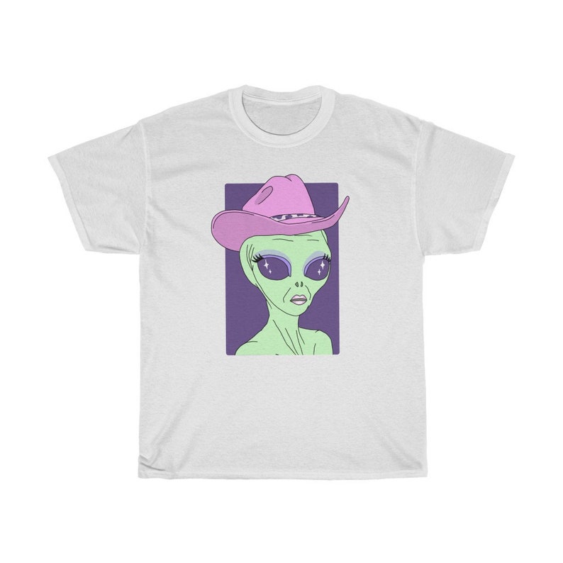 May include: A white t-shirt with a green alien wearing a pink cowboy hat. The alien has large, sparkling eyes and a purple background.