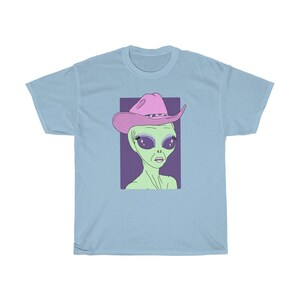 May include: A light blue t-shirt with a graphic of a green alien wearing a pink cowboy hat with sparkly eyes. The alien has a purple background.