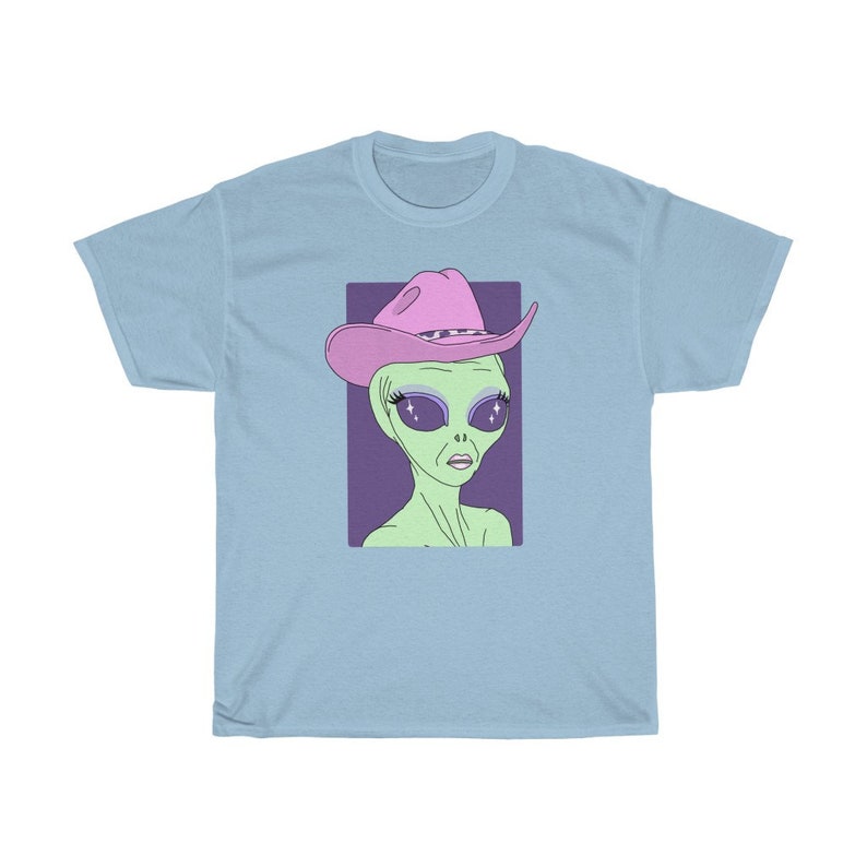 May include: A light blue t-shirt with a graphic of a green alien wearing a pink cowboy hat with sparkly eyes. The alien has a purple background.