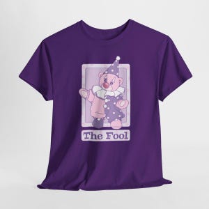 May include: A deep purple short-sleeve t-shirt featuring a graphic of a pink teddy bear dressed as a clown with a ruffled collar and a star-patterned hat. The design is framed in a light purple rectangle with the text The Fool printed below.