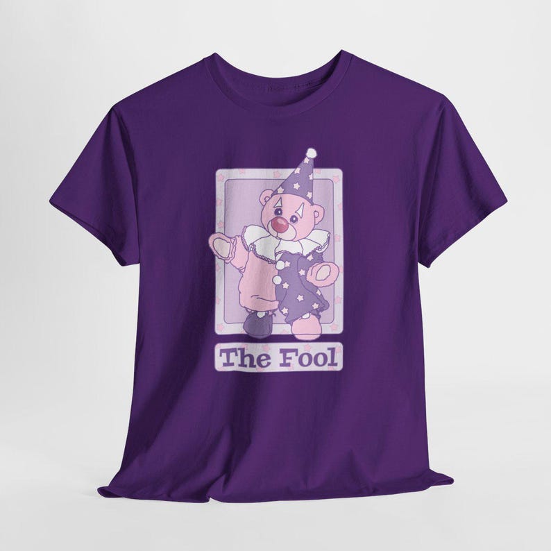 May include: A deep purple short-sleeve t-shirt featuring a graphic of a pink teddy bear dressed as a clown with a ruffled collar and a star-patterned hat. The design is framed in a light purple rectangle with the text The Fool printed below.