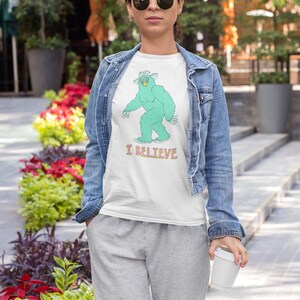 May include: A white t-shirt with a green monster graphic and the text "I BELIEVE". The person wearing the shirt is also wearing a denim jacket and grey sweatpants.