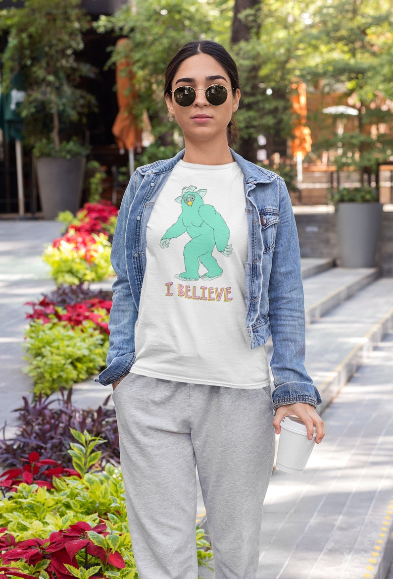 May include: A white t-shirt with a green monster graphic and the text "I BELIEVE". The person wearing the shirt is also wearing a denim jacket and grey sweatpants.