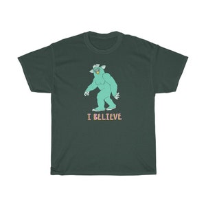 May include: A dark green t-shirt with a cartoon illustration of a green monster with a crown and the text "I BELIEVE" in pink and light blue.