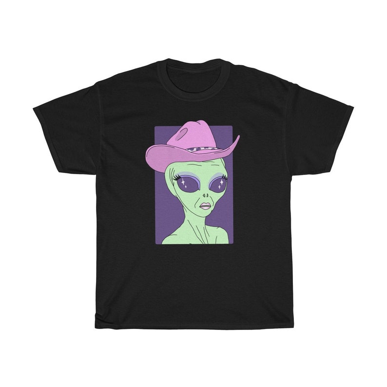 May include: A black t-shirt with a graphic of an alien wearing a pink cowboy hat. The alien has green skin, large eyes with glitter, and a serious expression.