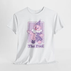 May include: A white short-sleeve t-shirt featuring a graphic of a teddy bear dressed as a clown in a purple and pink outfit with a ruffled collar and a star-patterned hat. Below the bear is a rectangular box containing the text The Fool.