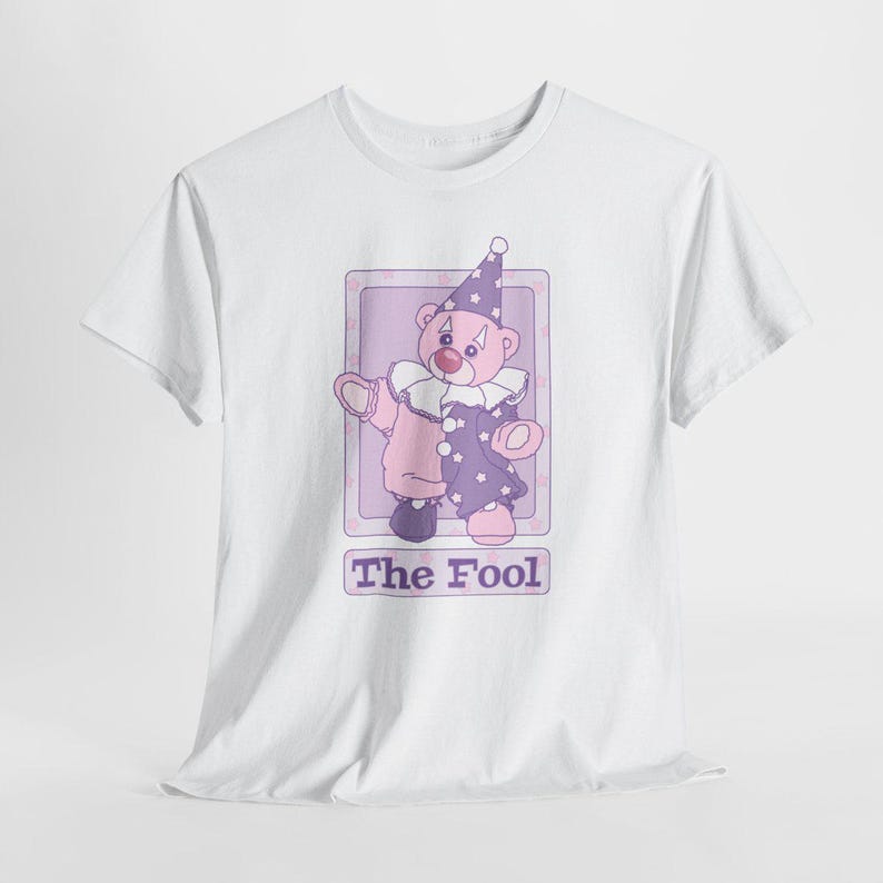 May include: A white short-sleeve t-shirt featuring a graphic of a teddy bear dressed as a clown in a purple and pink outfit with a ruffled collar and a star-patterned hat. Below the bear is a rectangular box containing the text The Fool.