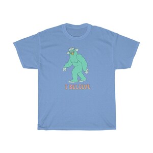 May include: A light blue t-shirt with a green cartoon monster walking and the text "I BELIEVE" in pink.