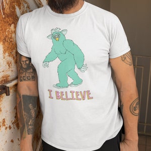 May include: White t-shirt with a green cartoon monster wearing glasses and the text "I BELIEVE" in pink and yellow letters.