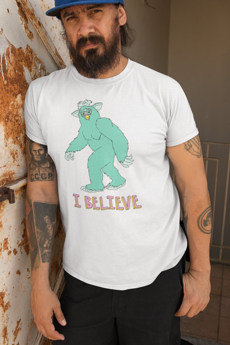 May include: White t-shirt with a green cartoon monster wearing glasses and the text "I BELIEVE" in pink and yellow letters.
