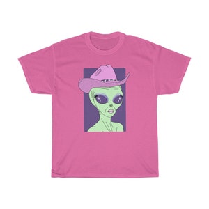 May include: A pink t-shirt with a graphic of a green alien wearing a pink cowboy hat with a purple band. The alien has large, sparkling eyes.