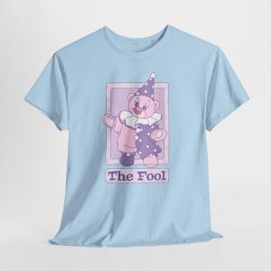 May include: A light blue t-shirt featuring a graphic of a teddy bear dressed as a clown in a purple and pink outfit with a matching hat and ruffled collar. The bear is inside a rectangular frame with the text The Fool printed below it.