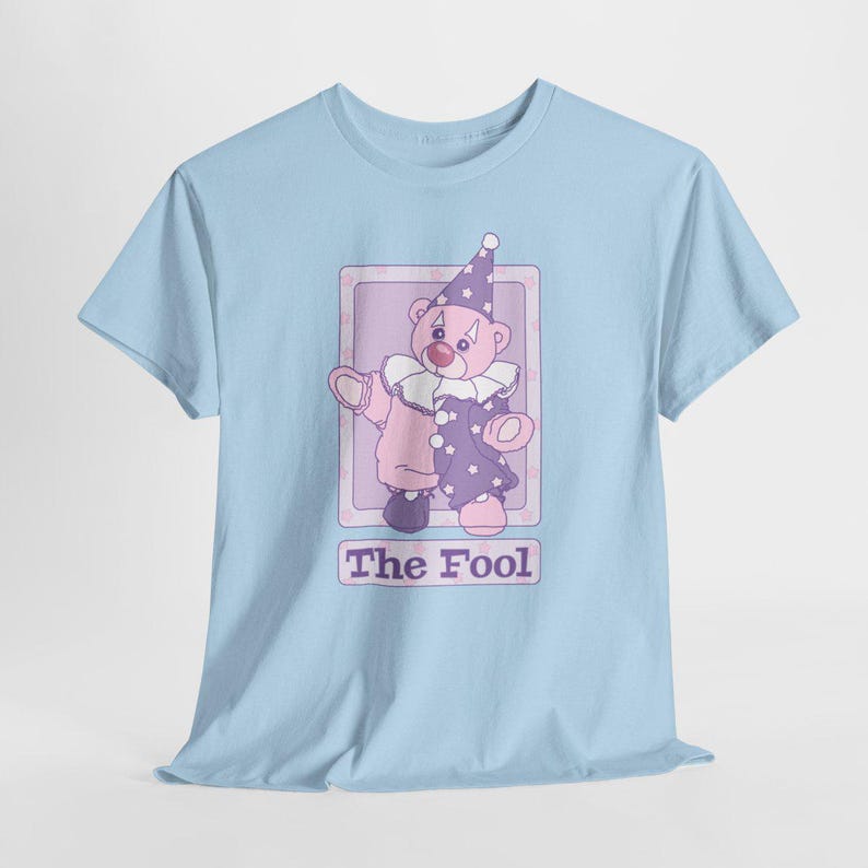 May include: A light blue t-shirt featuring a graphic of a teddy bear dressed as a clown in a purple and pink outfit with a matching hat and ruffled collar. The bear is inside a rectangular frame with the text The Fool printed below it.