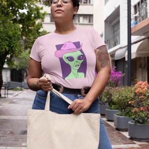 May include: A pink t-shirt with a graphic of an alien wearing a pink cowboy hat. The alien has green skin, large eyes, and a serious expression. The t-shirt is being worn by a person with short, curly hair and glasses. The person is holding a white tote bag.