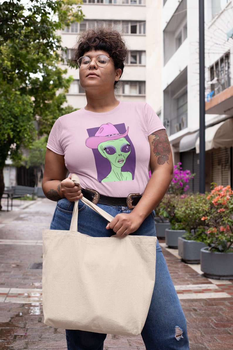 May include: A pink t-shirt with a graphic of an alien wearing a pink cowboy hat. The alien has green skin, large eyes, and a serious expression. The t-shirt is being worn by a person with short, curly hair and glasses. The person is holding a white tote bag.