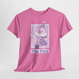 May include: A bright pink t-shirt featuring a graphic of a teddy bear dressed as a clown in a purple and pink outfit with a matching hat. The bear is inside a rectangular frame with the text The Fool printed in purple letters below the image.