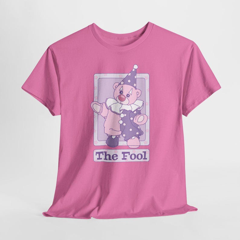 May include: A bright pink t-shirt featuring a graphic of a teddy bear dressed as a clown in a purple and pink outfit with a matching hat. The bear is inside a rectangular frame with the text The Fool printed in purple letters below the image.