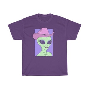 May include: A purple t-shirt with a graphic of an alien wearing a pink cowboy hat with sparkly eyes. The alien is green and has a light purple background.