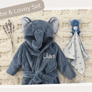 A blue elephant-shaped robe with the name "Liam" printed on it, along with a blue elephant-shaped blanket and a stuffed elephant toy, all arranged on a white knitted blanket.