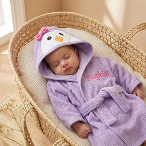 May include: A lavender hooded bathrobe featuring a penguin face on the hood with a pink bow, white accents, and an orange beak. The robe is made of soft terry cloth, includes a matching tie belt, and has the name Sophia embroidered in pink.
