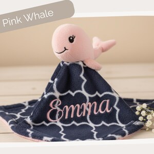 A pink plush toy of a whale is sitting on a blue and white patterned blanket with the name "Emma" embroidered on it.
