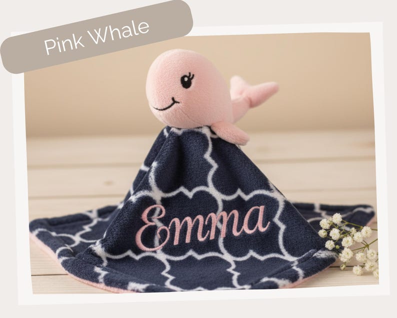 A pink plush toy of a whale is sitting on a blue and white patterned blanket with the name "Emma" embroidered on it.