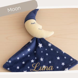 A plush toy of a crescent moon with a blue blanket featuring white stars and the name "Luna" printed on it.