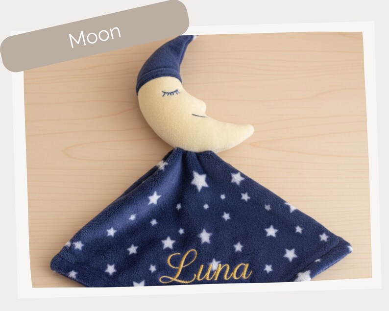A plush toy of a crescent moon with a blue blanket featuring white stars and the name "Luna" printed on it.