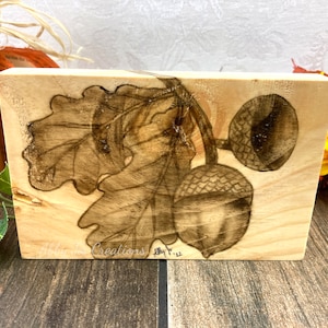 May include: A wood block with a woodburned image of oak leaves and acorns. The block is signed "Ally '22".