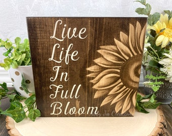 May include: A wooden sign with a brown background and white text that reads "Live Life In Full Bloom". A brown and beige sunflower is painted on the right side of the sign.