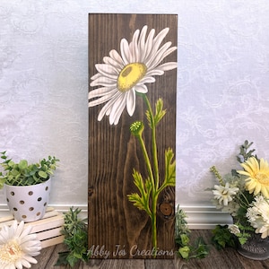 May include: A hand-painted wooden sign featuring a white daisy with a yellow center on a dark brown wood background. The sign is titled "Abby Jo's Creations".