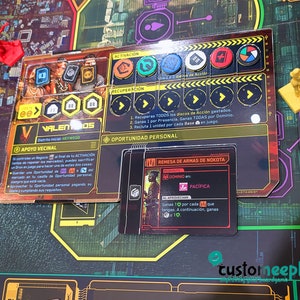 May include: A close-up of a board game card with a black and red border. The card features a character with a black and white patterned shirt and a black and white patterned hat. The card text reads "Remesa de Armas de Nokota" and "Dominio Pac&iacute;fica".