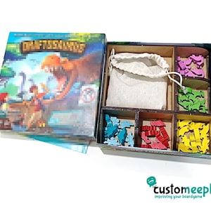May include: A board game box set titled "Draftosaurus" displays a dinosaur illustration. Inside, wooden game pieces in blue, red, yellow, green, and purple are arranged in compartments, alongside a beige drawstring bag. The "customeeple" logo is visible.