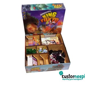 May include: A wooden board game organiser with several compartments, filled with game pieces, cards, and tokens. The box features the game title "King of Tokyo" and a colourful graphic design. The organiser is designed to hold and organise the game components.