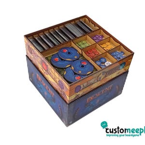 May include: A wooden board game organiser with various compartments. It holds game pieces, cards, and tokens. The box features a blue and brown design, with the word "DESCENT" printed on the side. The organizer is designed to keep the game components tidy.