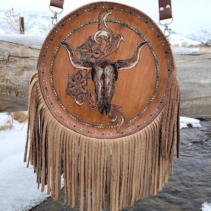 May include: A round brown leather purse with a fringe border. The purse has a skull with horns and floral designs painted on it. The purse has a brown leather strap.