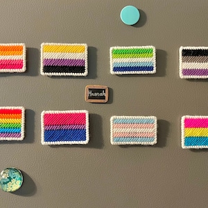 May include: A collection of 10 small, colorful, woven flags on a gray wall. The flags represent different pride flags, including the transgender, genderqueer, lesbian, gay, bisexual, and pansexual flags. The flags are arranged in two rows of five. There is a small sign that says "Hannah" on the wall.