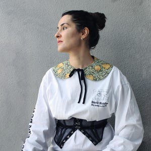 Könnte beinhalten: Weißes Langarmshirt mit dekorativem Blumenkragen in Gelb und Grün sowie einer schwarzen Schleife. Das Shirt trägt den Text "Devil Hunter My name is Perry" und eine Grafik. Ein schwarzes Korsett wird über dem Shirt getragen.