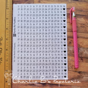 May include: A white sticker sheet featuring rows of black printed numbers from 1 to 31, each ending with a black heart icon. The sheet includes the text Charlie B's Papeterie and 365 Font: Benilla Calligraphy, measured by a 7-inch wooden ruler.