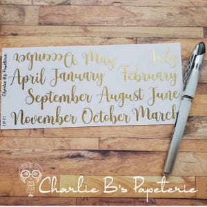 Puede incluir: Una hoja de pegatinas blanca con letras doradas de los meses del año. Un bolígrafo plateado descansa sobre la superficie de madera. La hoja de pegatinas tiene el texto "Charlie B's Papeterie" y "LM 01".