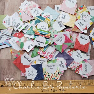May include: A collection of small, folded paper envelopes in various patterns and colors, each containing a white note with printed text such as 'You shine', 'Love you lots', 'Bloom', 'Be awesome', 'Just for you', 'Fabulous', and 'Miss your face'.