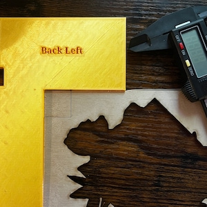 May include: A gold-colored plastic corner piece with the words "Back Left" printed on it. The corner piece is sitting on a wooden surface next to a piece of wood with a floral design cut out.