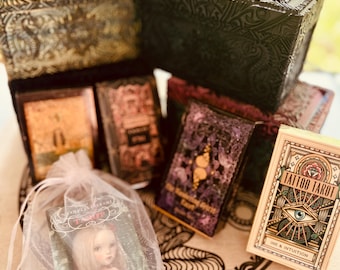 May include: A collection of tarot decks, including a deck with a doll illustration, a deck with a purple and gold design, and a deck with a tattoo-inspired design. The decks are in their boxes, which are made of wood and have intricate carvings.