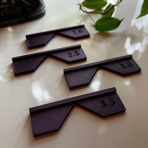 May include: Four purple plastic rulers with different measurements: 2.0, 2.5, 3.0, and 3.5. The rulers are arranged in a row on a white surface.