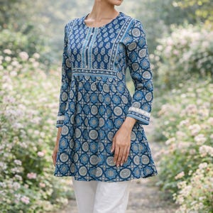 May include: A woman wearing a long-sleeved, blue tunic with a white and gold pattern, paired with white pants. The tunic has a round neckline and a flared hem. The sleeves are long and end with a white lace trim. The woman is standing on a path surrounded by greenery.